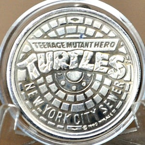 May include: A silver coin featuring the Teenage Mutant Ninja Turtles logo. The coin is round, with the words "Teenage Mutant Hero Turtles", "Turtles", "New York City Sewer" and the year 1990. The coin is encased in a clear plastic holder.
