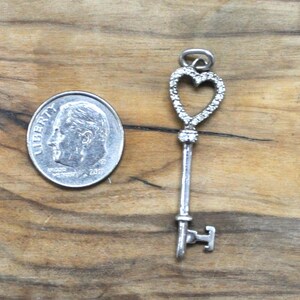 May include: A silver key charm with a heart-shaped design. The heart is outlined with small, clear stones. The key is about the size of a dime.