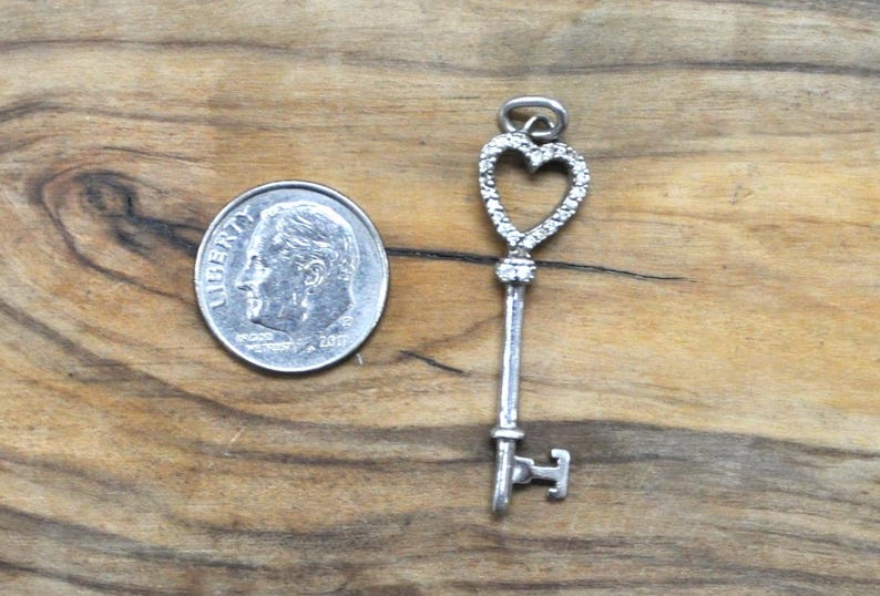 May include: A silver key charm with a heart-shaped design. The heart is outlined with small, clear stones. The key is about the size of a dime.