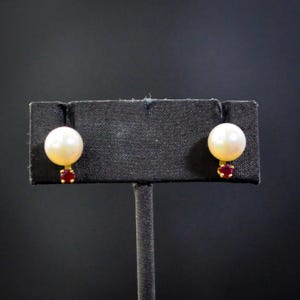 May include: A pair of stud earrings featuring a large, round, white pearl set above a small, red gemstone. The earrings have a gold-colored setting and are displayed on a black jewelry stand against a dark background. These earrings are a classic jewelry item.