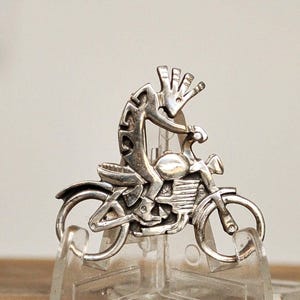 May include: A silver-coloured metal pin showing a stylised figure riding a motorcycle. The figure has an elongated body and a headdress. The motorcycle has detailed wheels and engine components. The pin is displayed on a clear stand.