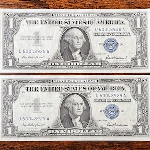 Pode incluir: Duas notas antigas de certificado de prata de um dólar. Cada nota apresenta um retrato de George Washington e o texto "ONE DOLLAR" em uma moldura decorativa. As notas são da Série de 1957.