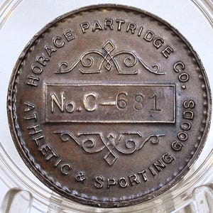 Peut inclure: Un jeton métallique circulaire vintage avec le texte "HORACE PARTRIDGE CO. ATHLETIC & SPORTING GOODS" et "No. C-681" dans un rectangle. Le jeton est dans un support en plastique transparent.