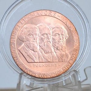 May include: A copper-coloured United States commemorative medal featuring three men with beards. The medal is inscribed with "RUSSELL * MAJORS * WADDELL" and "UNITED STATES COMMEMORATIVE MEDAL FOUNDERS". The medal is encased in a clear plastic holder.