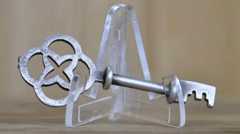 May include: A silver key with a decorative, ornate head in the shape of a four-leaf clover. The key is displayed on a clear plastic stand.