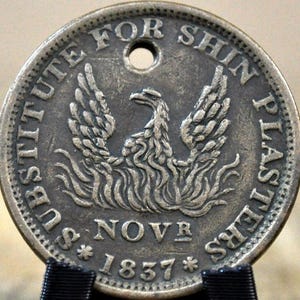 May include: A circular, antique coin with the text "SUBSTITUTE FOR SHIN PLASTERS" and "NOVR 1837". The coin features a detailed image of a bird with outstretched wings. The coin has a hole at the top.