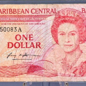 Puede incluir: Un billete de un dólar del Banco Central del Caribe Oriental. El billete presenta un retrato de una mujer con corona y collar. El texto "ONE DOLLAR" se muestra de forma destacada, junto con el nombre del banco y la denominación.