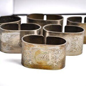May include: A set of six oval-shaped silver-toned metal napkin rings, each featuring an intricate engraved floral pattern and the monogrammed letters HS centered on the front surface, arranged in a cluster on a plain white surface.