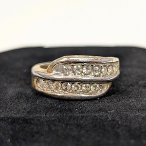 May include: A silver-toned ring with a gold band and two rows of clear, sparkling stones. The ring has a unique, overlapping design, and is set against a dark background.