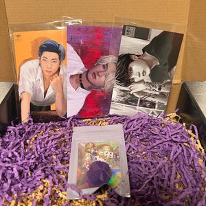 Puede incluir: Una caja llena de papel de seda morado que contiene tres tarjetas ilustradas con diferentes hombres, y una peque&ntilde;a bolsa transparente con un llavero de pomp&oacute;n morado con abalorios dorados y cuentas de letras.