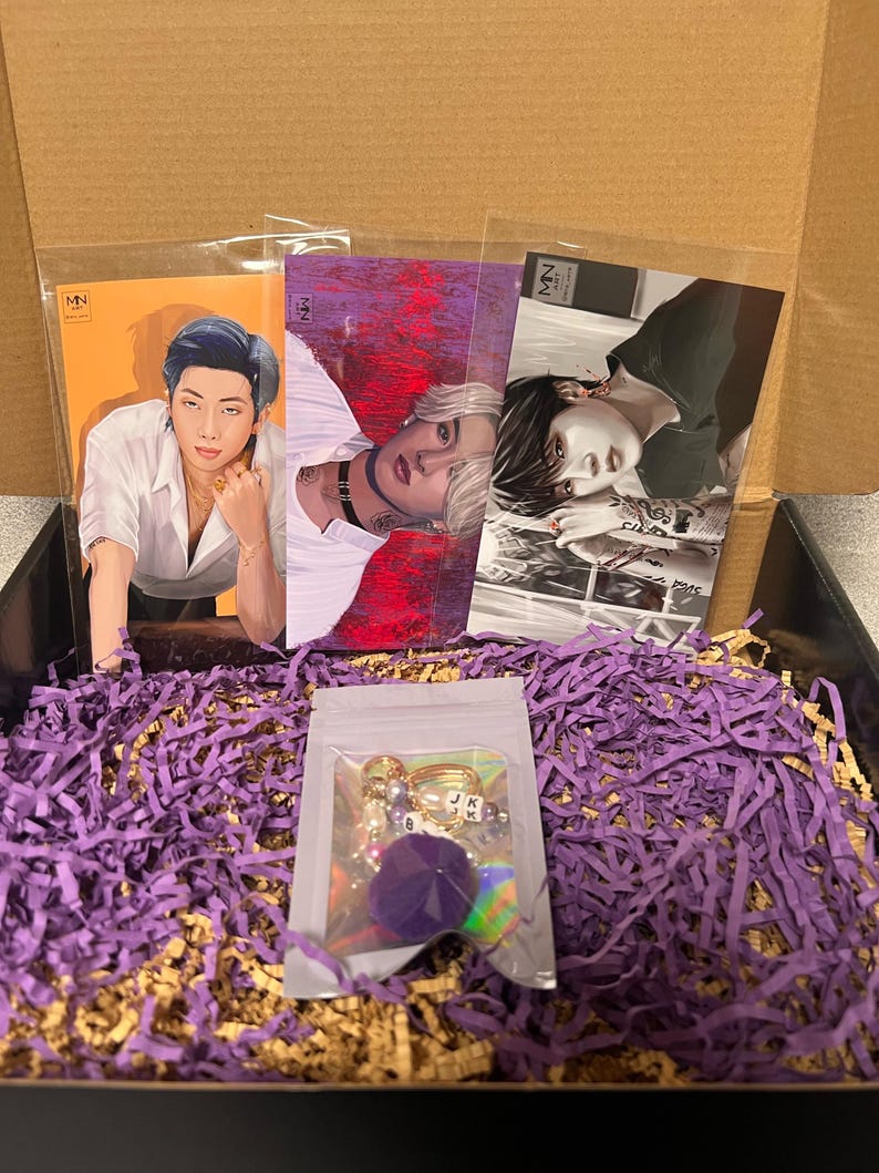 Puede incluir: Una caja llena de papel de seda morado que contiene tres tarjetas ilustradas con diferentes hombres, y una peque&ntilde;a bolsa transparente con un llavero de pomp&oacute;n morado con abalorios dorados y cuentas de letras.