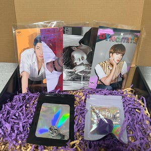 May include: A purple and white gift box filled with purple shredded paper. The box contains four photo cards featuring a K-pop band member, a silver charm necklace, and a purple charm bracelet with the letters "JK".