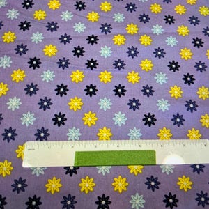 May include: A lavender fabric with a repeating pattern of yellow, light blue, dark blue, and black daisy-like flowers. A ruler with inch measurements is placed on the fabric.