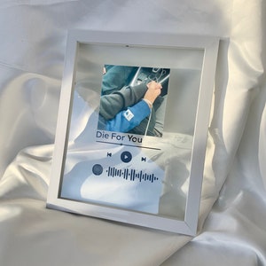 May include: A white framed print with a photograph of two people holding hands. The print includes the text "Die For You" and a music player interface with a waveform graphic. The frame is rectangular and rests on a white, silky surface.