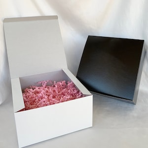 Peut inclure: Deux boîtes cadeaux, une blanche et une noire, sont présentées. La boîte blanche est ouverte, révélant du papier déchiqueté rose à l'intérieur. La boîte noire est fermée. Les deux boîtes ont une finition brillante, sur fond blanc.