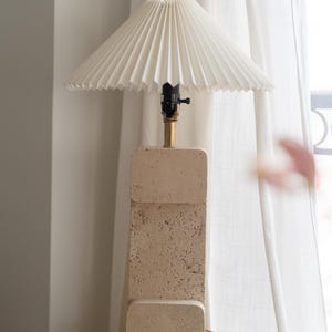 Puede incluir: Una l&aacute;mpara de mesa de piedra beige con una pantalla blanca plisada. La l&aacute;mpara est&aacute; sobre una base de piedra beige.