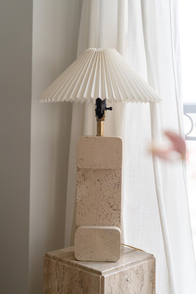 Puede incluir: Una l&aacute;mpara de mesa de piedra beige con una pantalla blanca plisada. La l&aacute;mpara est&aacute; sobre una base de piedra beige.