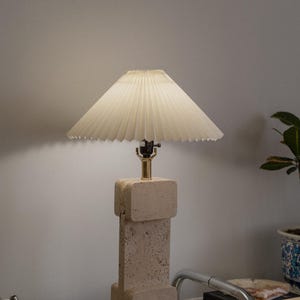 Puede incluir: Una l&aacute;mpara beige con una pantalla blanca plisada se encuentra sobre una superficie a cuadros. La base de la l&aacute;mpara est&aacute; hecha de un material texturizado que se asemeja a la piedra.