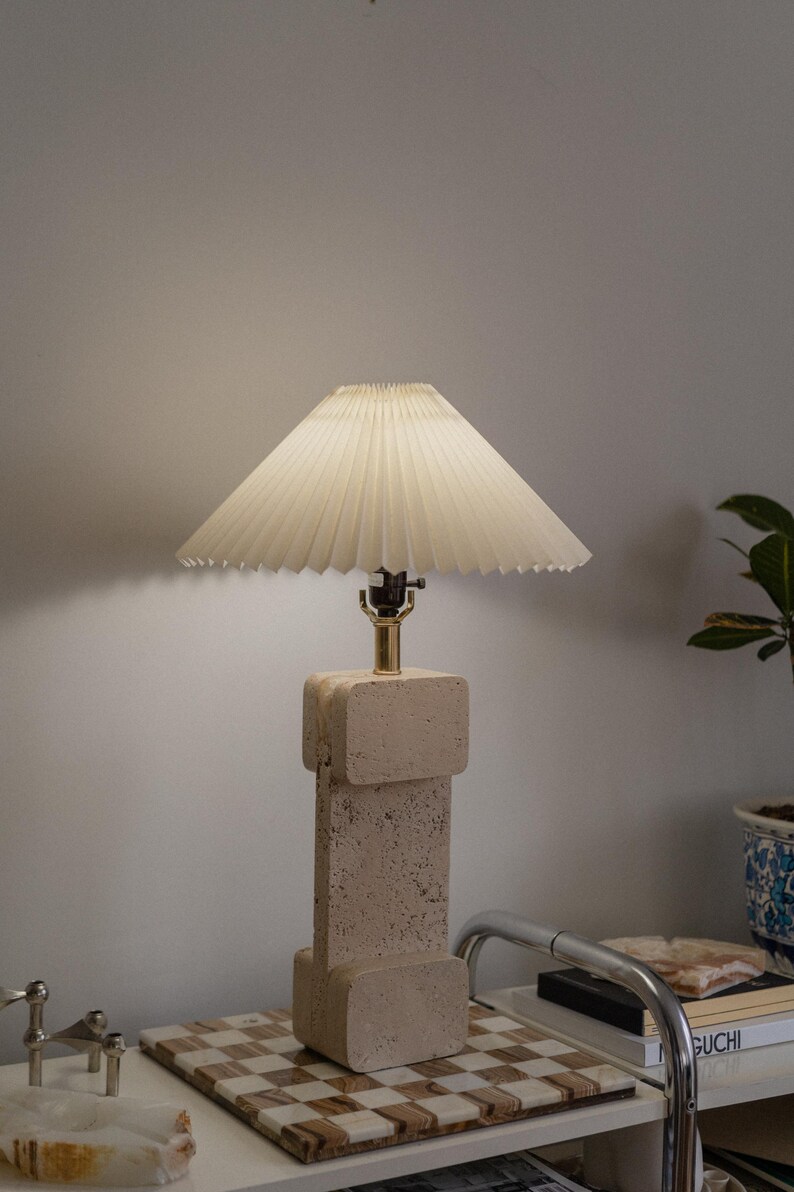Puede incluir: Una l&aacute;mpara beige con una pantalla blanca plisada se encuentra sobre una superficie a cuadros. La base de la l&aacute;mpara est&aacute; hecha de un material texturizado que se asemeja a la piedra.