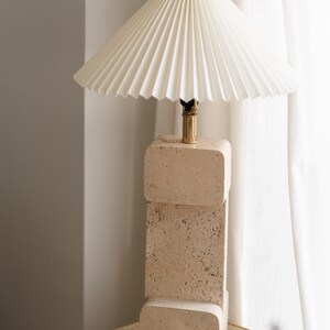 Puede incluir: Una l&aacute;mpara de mesa de travertino beige con una pantalla de l&aacute;mpara blanca plisada. La l&aacute;mpara est&aacute; sobre una base de travertino a juego.
