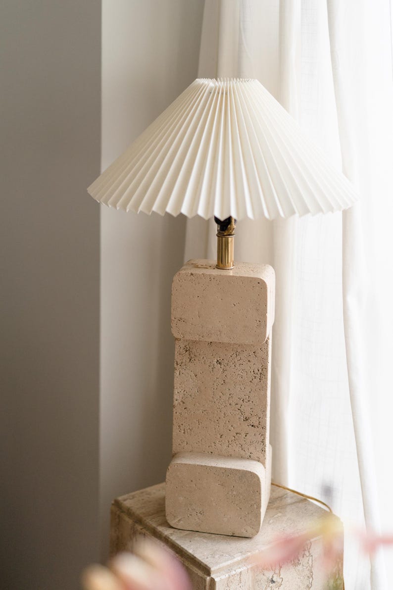 Puede incluir: Una l&aacute;mpara de mesa de travertino beige con una pantalla de l&aacute;mpara blanca plisada. La l&aacute;mpara est&aacute; sobre una base de travertino a juego.