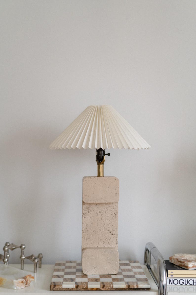 Puede incluir: Una l&aacute;mpara de mesa de piedra beige con una pantalla blanca plisada. La l&aacute;mpara est&aacute; sobre una superficie a cuadros con un libro y una bandeja al lado.