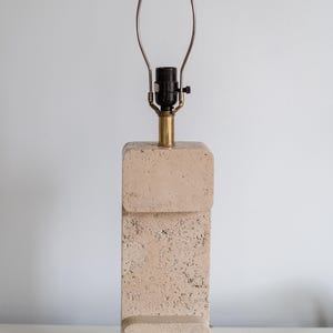 Puede incluir: Una l&aacute;mpara de mesa de travertino beige con base de lat&oacute;n y una pantalla de metal simple.