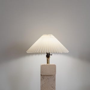 Puede incluir: Una l&aacute;mpara beige con una pantalla blanca plisada se encuentra sobre una superficie a cuadros. La base de la l&aacute;mpara est&aacute; hecha de un material texturizado, posiblemente piedra.