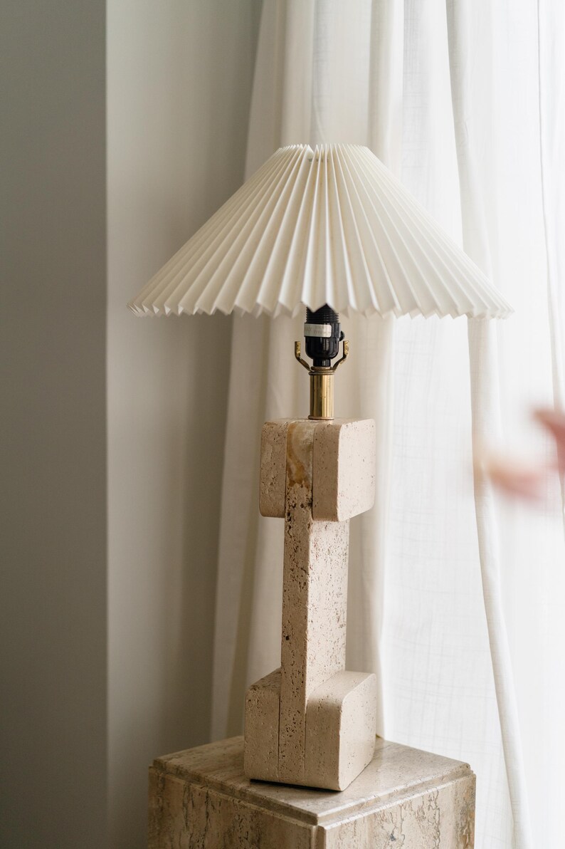 Puede incluir: Una pantalla de l&aacute;mpara blanca plisada se asienta sobre una base de l&aacute;mpara de piedra beige. La l&aacute;mpara est&aacute; sobre una mesa de piedra beige.