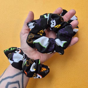 May include: Black scrunchies with a Halloween-themed print. The fabric features white ghosts, the word "BOO!", and the number "100!" in orange, purple, and green. The scrunchies are displayed against a yellow background.