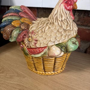 May include: A ceramic rooster-shaped lidded jar featuring a cream-colored body, a bright red comb, and a multi-colored tail with blue, green, and brown feathers. The base is shaped like a woven basket filled with sculpted fruit and flowers.
