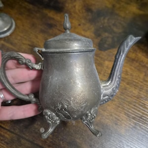 May include: A tarnished silver-toned metal teapot featuring a bulbous body, a hinged lid with a pointed finial, an ornate curved handle, and a long spout. The teapot stands on three decorative feet and displays visible surface wear and scratches.