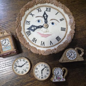 May include: A collection of five decorative clocks. The largest is a round wall clock with a human face, Roman numerals, and a brown ornate border. Four smaller clocks include a rectangular mantel clock, two round clocks, and a small arched clock with floral details.
