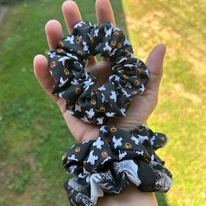 May include: Two black and white scrunchies with a Halloween theme. One scrunchie has a pattern of white ghosts and orange pumpkins on a black background. The other scrunchie has a black and white pattern of a spooky scene.