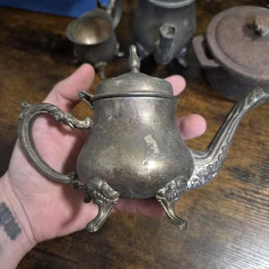May include: A tarnished silver-toned metal teapot featuring a curved spout, an ornate handle with scrollwork details, a removable lid with a pointed finial, and three decorative feet. The surface shows signs of age and oxidation.