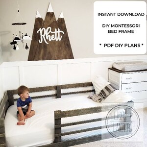 May include: DIY Montessori bed frame plans. A white wooden sign with the name "Rhett" and a mountain range design is hanging on the wall above a wooden bed frame with a white mattress and a child sitting on the bed. The bed frame has a safety rail on one side. The image also includes a dresser with three drawers and a white changing table.