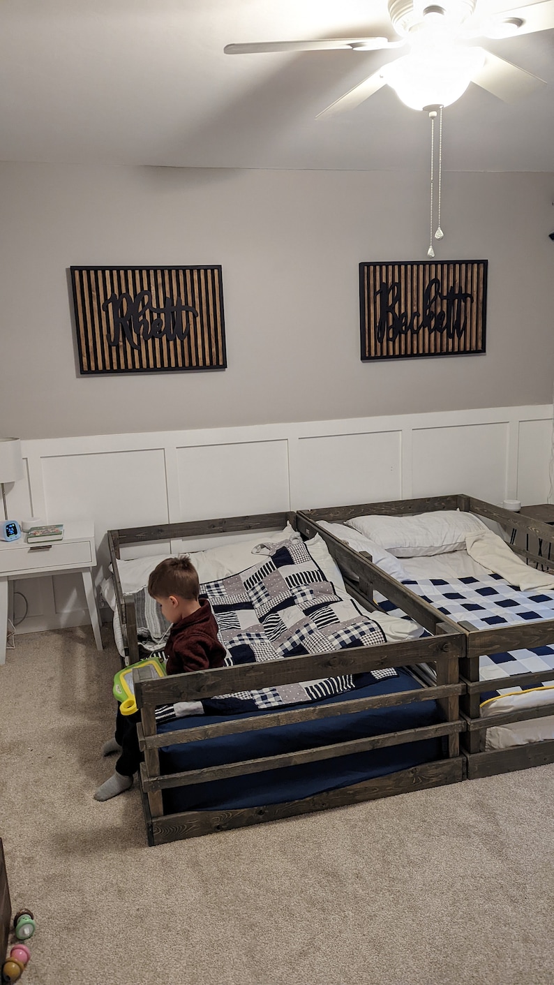 May include: Two wooden toddler beds with guard rails, one with a black and white checked quilt and a child sitting on it, and the other with a white pillow and a blue and white checked quilt. The beds are in a room with white walls and a ceiling fan.