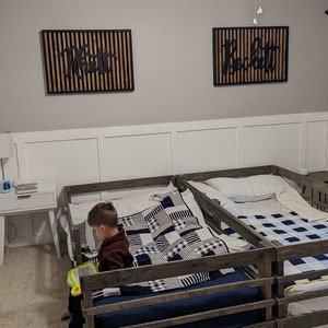 May include: Two wooden toddler beds with guard rails, one with a black and white checkered quilt and a child sitting on it, and the other with a white pillow and a blue and white checkered quilt. The beds are in a room with white walls and a ceiling fan.