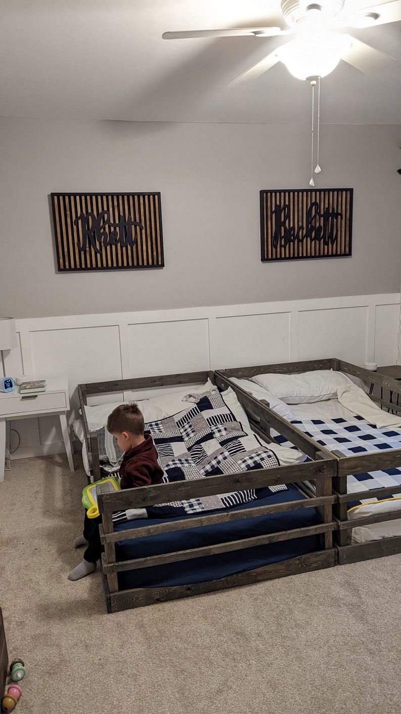 May include: Two wooden toddler beds with guard rails, one with a black and white checkered quilt and a child sitting on it, and the other with a white pillow and a blue and white checkered quilt. The beds are in a room with white walls and a ceiling fan.