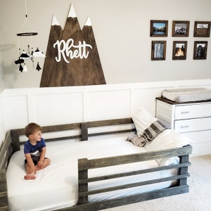 May include: A toddler sits on a white bed with a wooden bed rail. The bed is against a white wall with a wooden mountain range wall decor with the name "Rhett" written on it. There are framed pictures on the wall to the right of the bed.
