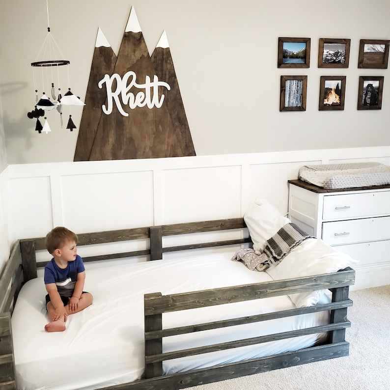 May include: A toddler sits on a white bed with a wooden bed rail. The bed is against a white wall with a wooden mountain range wall decor with the name "Rhett" written on it. There are framed pictures on the wall to the right of the bed.