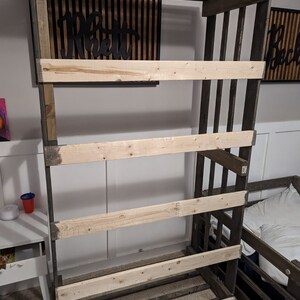 May include: A wooden shelf unit with four shelves, constructed from dark stained wood with light wood shelves. The shelf unit is standing in a room with a white wall and a light wood floor.