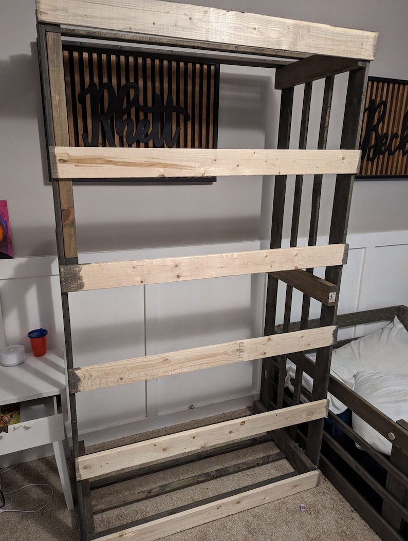 May include: A wooden shelf unit with four shelves, constructed from dark stained wood with light wood shelves. The shelf unit is standing in a room with a white wall and a light wood floor.