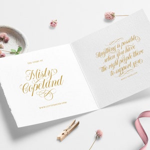 May include: A white card with gold lettering that reads "The Story of Misty Copeland" on the front and "Anything is possible when you have the right people there to support you" on the back. The card is surrounded by pink flowers and a pink ribbon.