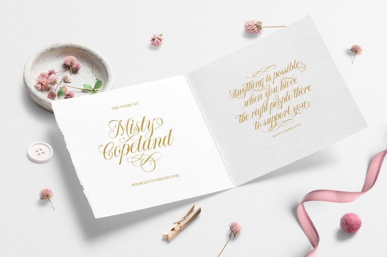 May include: A white card with gold lettering that reads "The Story of Misty Copeland" on the front and "Anything is possible when you have the right people there to support you" on the back. The card is surrounded by pink flowers and a pink ribbon.