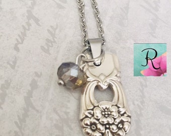 May include: A silver necklace with a vintage spoon charm and a dark gray crystal bead. The spoon charm has a floral design.