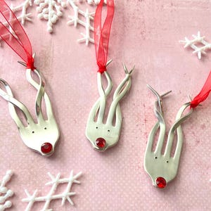 May include: Three silver reindeer ornaments made from forks, each with a red gem nose and red ribbon for hanging. The ornaments are on a pink background with white snowflake decorations.