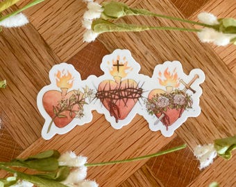 May include: Three stylized hearts with flames, thorns, and a sword, representing the Sacred Hearts of Jesus and Mary. The hearts are drawn in a vintage style with watercolor-like details.