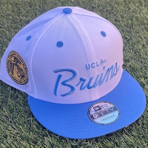 May include: A white and blue snapback hat with the text "UCLA Bruins" embroidered in blue script. The hat features a blue brim and a patch on the side. The hat has a 9FIFTY Snapback sticker.