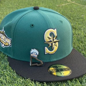 May include: A green baseball cap with a black brim. The cap features a gold and white logo of a team, a patch with text, and a pin of a cartoon character. The cap size is 7 1/4.